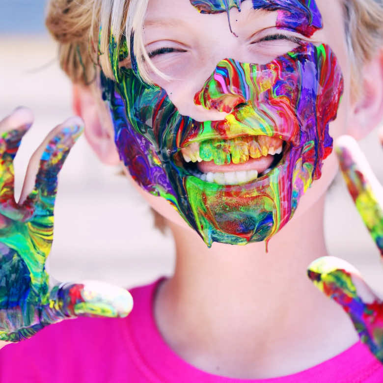 A young boy, smiling, with multi-coloured paint all over his face and hands